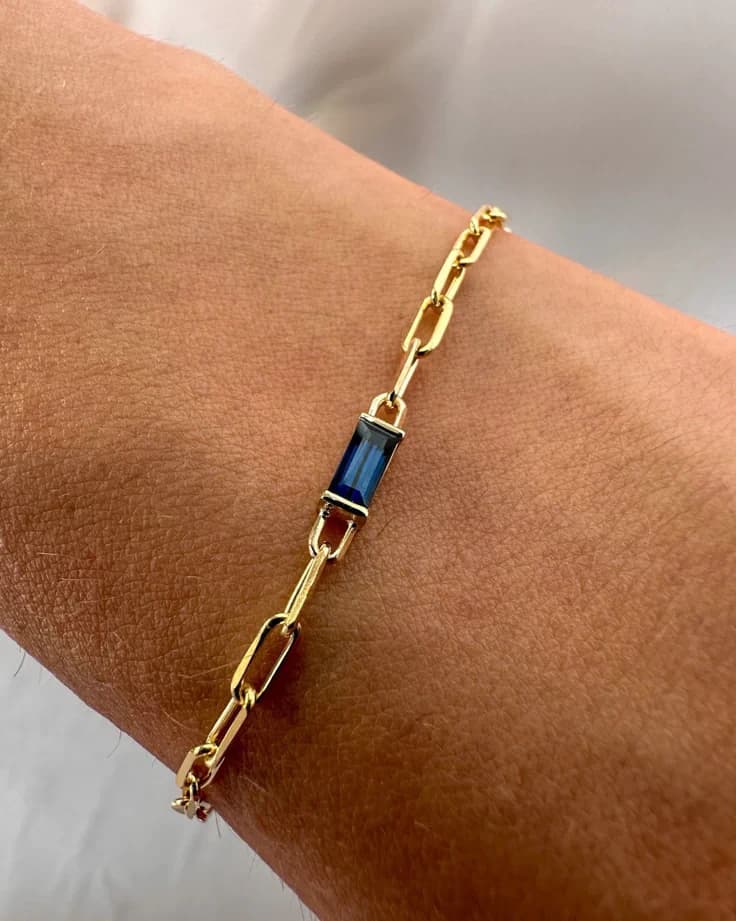 Gold paperclip-link bracelet with a blue baguette stone, worn on the wrist